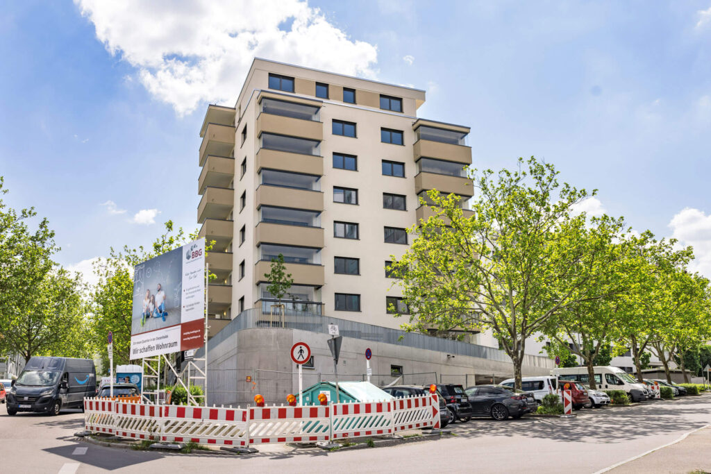 Modernes Mehrfamilienhaus mit Balkonen an einer Straßenecke umgeben von Bäumen. Im Vordergrund ein Baustellenzaun