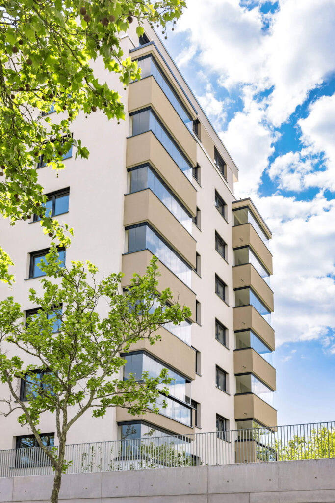 Ausschnitt eines modernen Mehrfamilienhauses mit Balkonen, im Vordergrund Blätter und eine Betonmauer