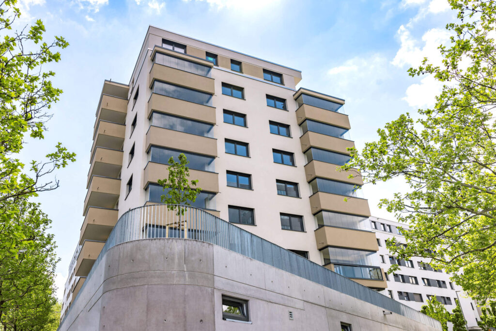 Modernes Mehrfamilienhaus aus der Froschperspektive umgeben von Bäumen, im Vordergrund eine Betonmauer. Im Hintergrund weitere Wohngebäude.