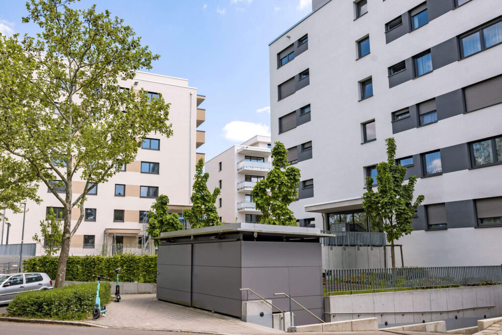 Moderne Mehrfamilienhäuser mit Bäumen und einem grauen Nebengebäude im Vordergrund bei blauem Himmel