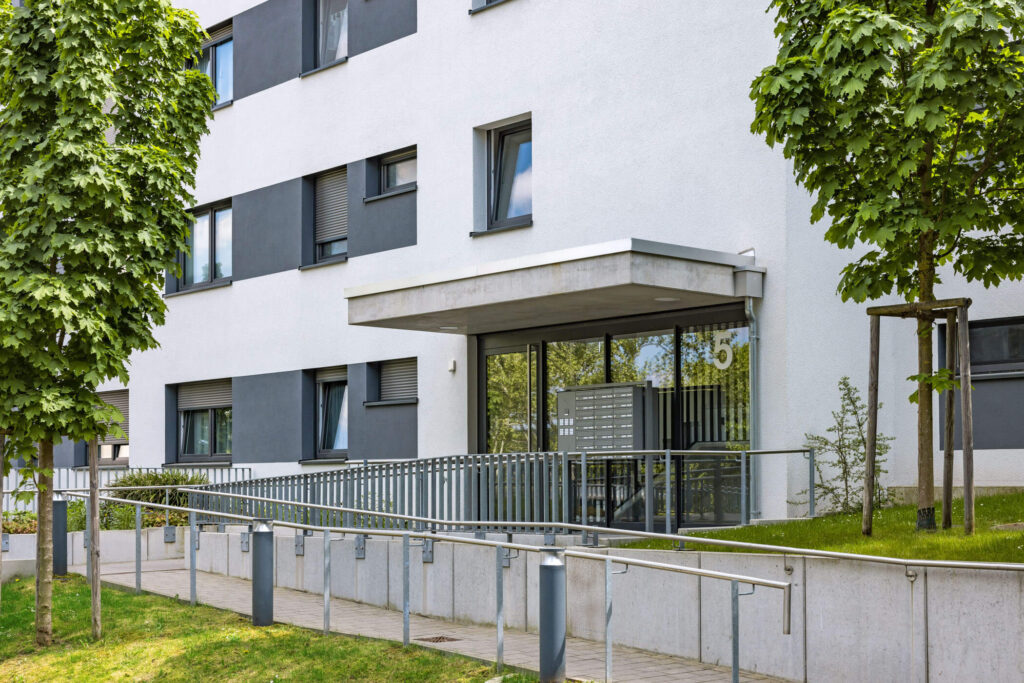 Eingangsbereich eines modernen Wohnhauses mit Rampe, Briefkästen und Hausnummer 5, umgeben von Bäumen und Rasen