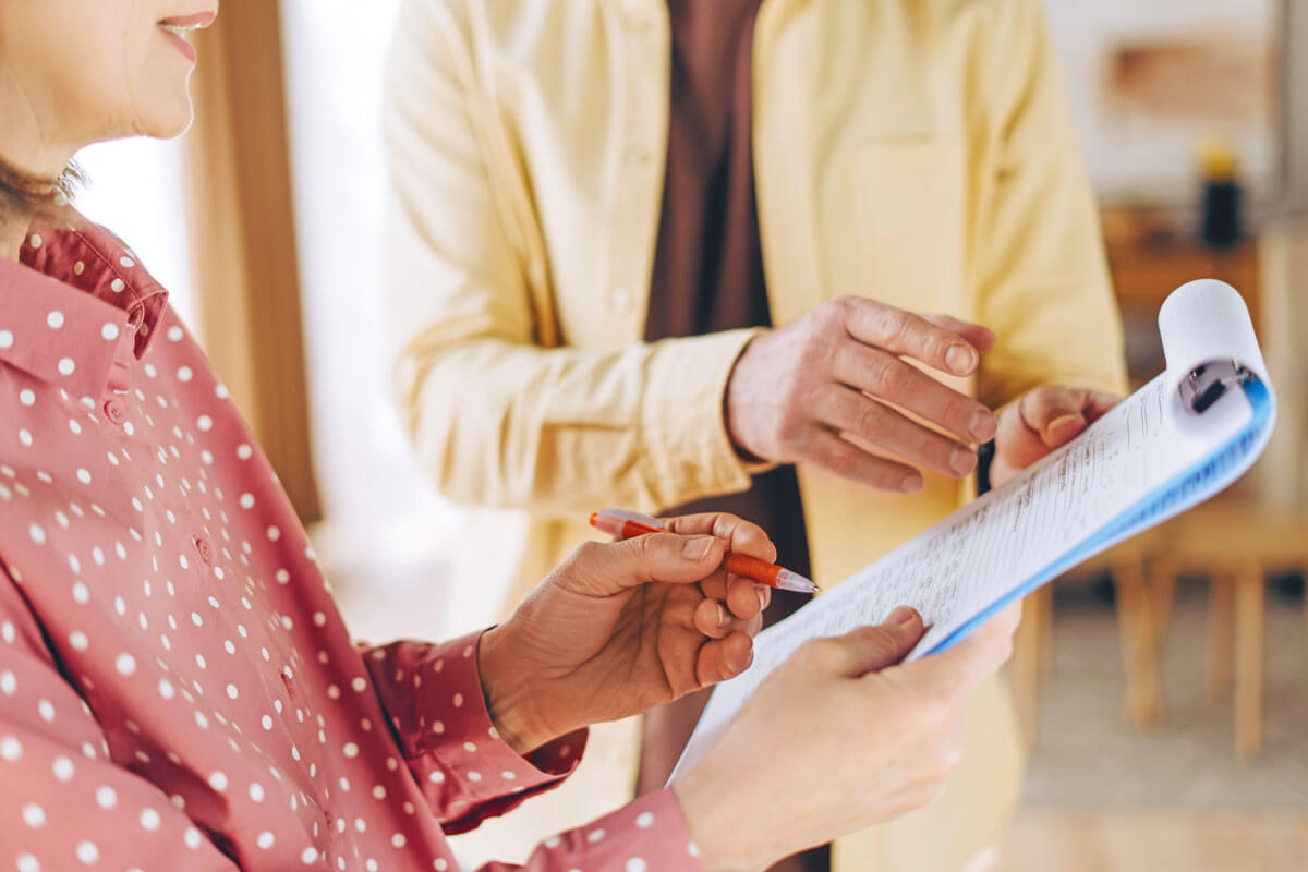 Nahaufnahme einer Frau mittleren Alters, die Zeilen auf einer Checkliste abhakt, neben ihr ein jüngerer Mann