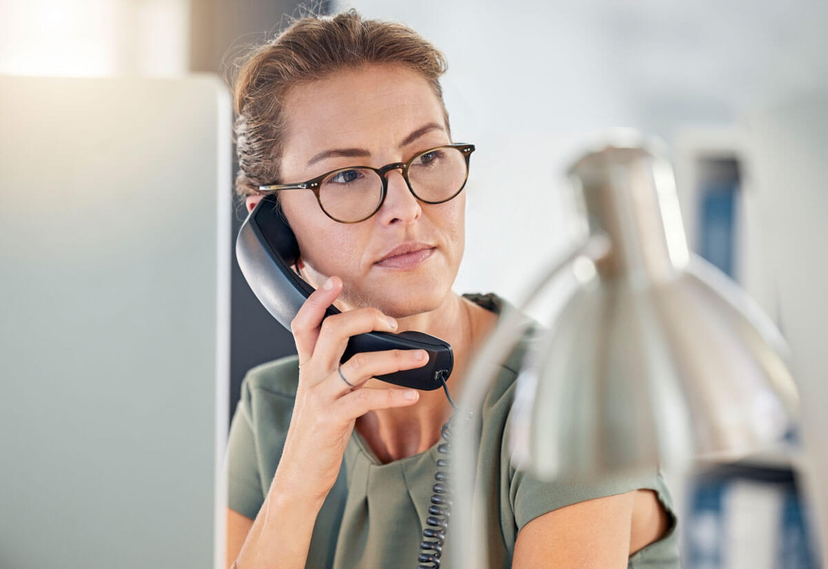 Eine ernste Frau am Telefon, im Vordergrund unscharf Schreibtischgegenstände