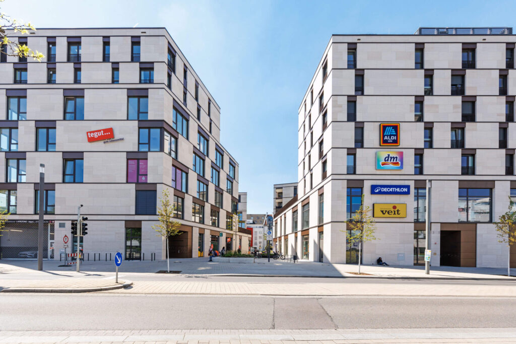 Frontalansicht modernes Wohn- und Gewerbequartiers durch zwei Hauptgebäude begrenzt, mit prominenten Schildern aller Geschäfte am Gebäude und der anliegenden Straße im Vordergrund