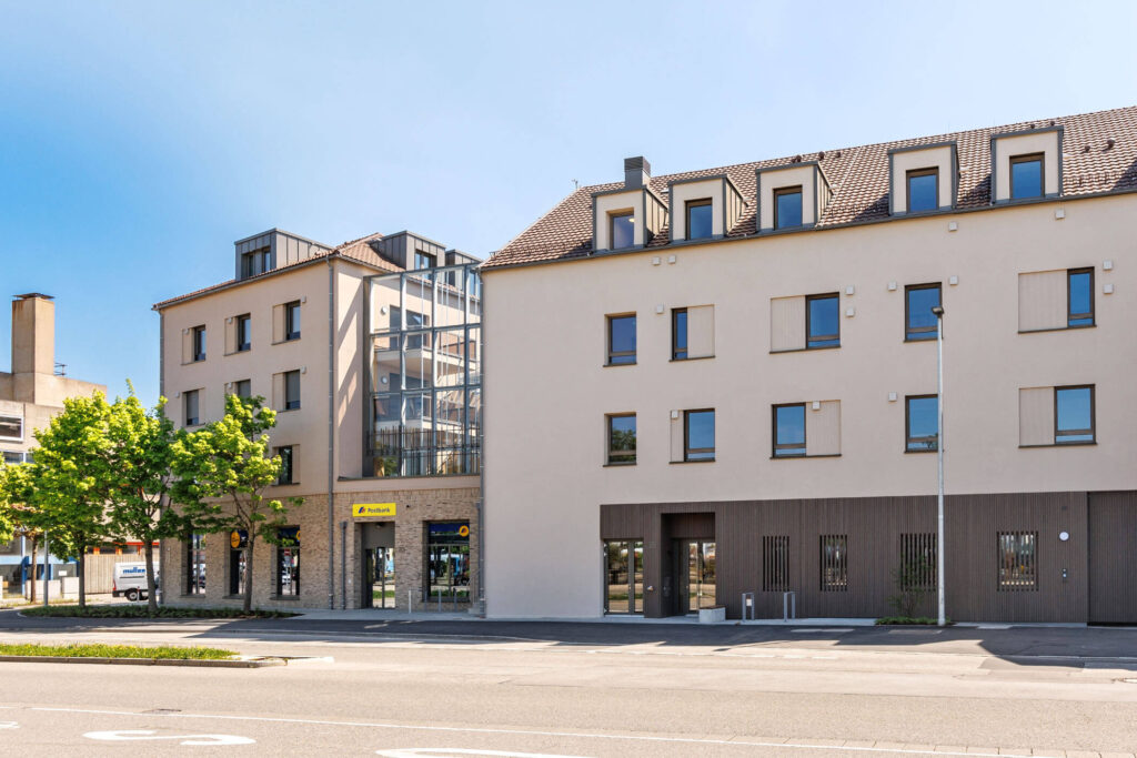 Würdevoller, beigefarbener Gebäudekomplex aus zwei Häusern mit vielen Fenstern, Erkerfenstern und teilweise verglaster Fassade. Das linke Gebäude hat ein wundervoll gemauertes Erdgeschoss, das rechte dort eine Holzvertäfelung. Vor dem Eingangsbereich Farhrradständer und Sitzbänke, davor Bäume und eine Straße.