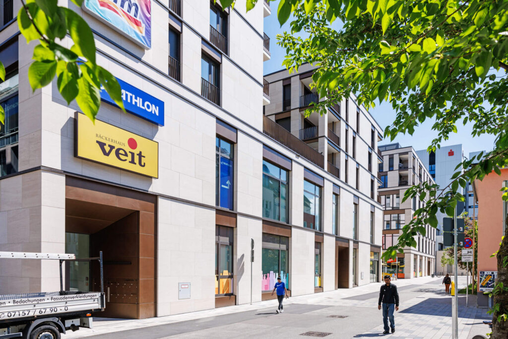 Bildausschnitt eines modernen Wohn- und Gewerbequartier mit Schildern einiger Läden an der Wand, davor laufen Menschen. Im Vordergrund Blätter.