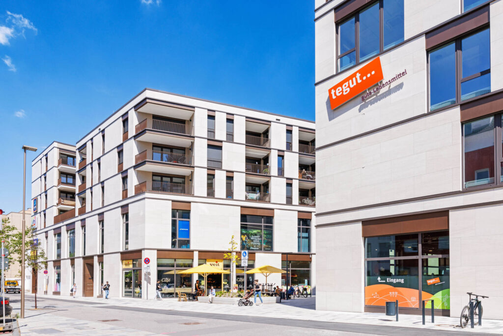 Ein modernes Wohn- und Gewerbequartier begrenzt durch die beiden Hauptgebäude, mit Menschen und Sonnenschirmen vor einer Bäckerei