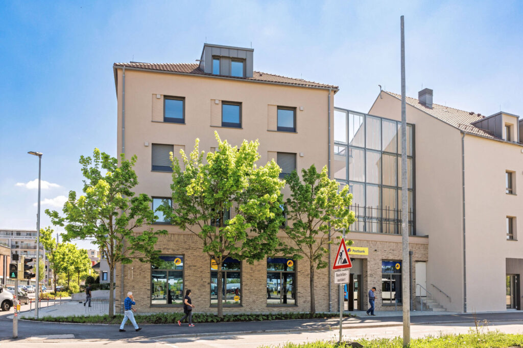 Würdevoller, beigefarbener Gebäudekomplex aus zwei Häusern mit vielen Fenstern, Erkerfenstern und teilweise verglaster Fassade. Das linke Gebäude mit einem wundervoll gemauerten Erdgeschoss. Vor dem Eingangsbereich Fahrradständer und Autostellflächen, davor Bäume, einige Fußgänger und eine Straße.