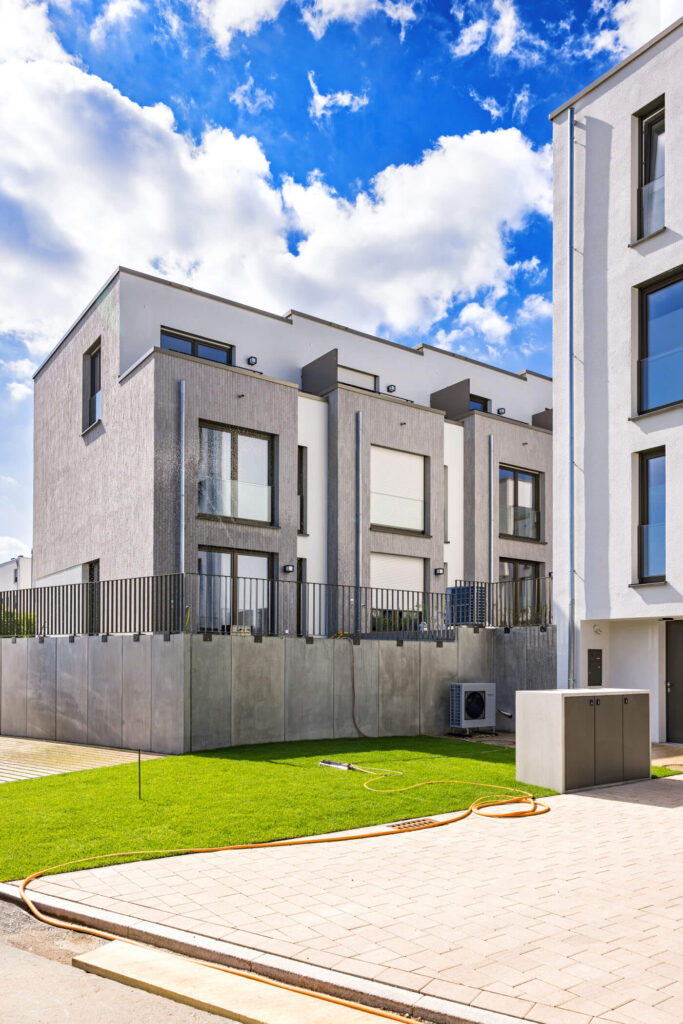 Moderne, wunderschöne weiße Reihenhäuser mit grauen Zierfassaden in einer eleganten Wohngegend hinter einer betonierten Abgrenzung, davor etwas gepflegtes Grün und die Einfahrt eines weiteren Wohnhauses im Quartier, das am rechten Rand erkennbar ist