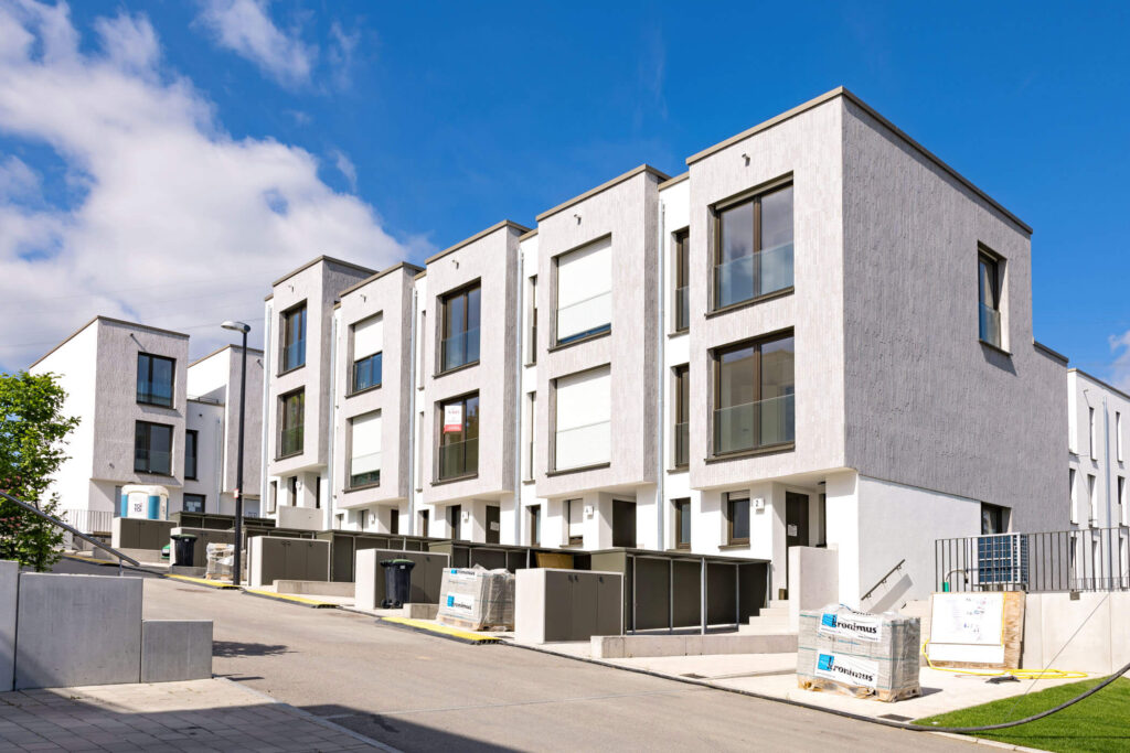 Fünf moderne, wunderschöne weiße Reihenhäuser mit grauen Zierfassaden an einer leicht ansteigenden Straße umgeben von Baumaterialien auf Paletten, im Hintergrund weitere Wohnhäuser des Quartiers.