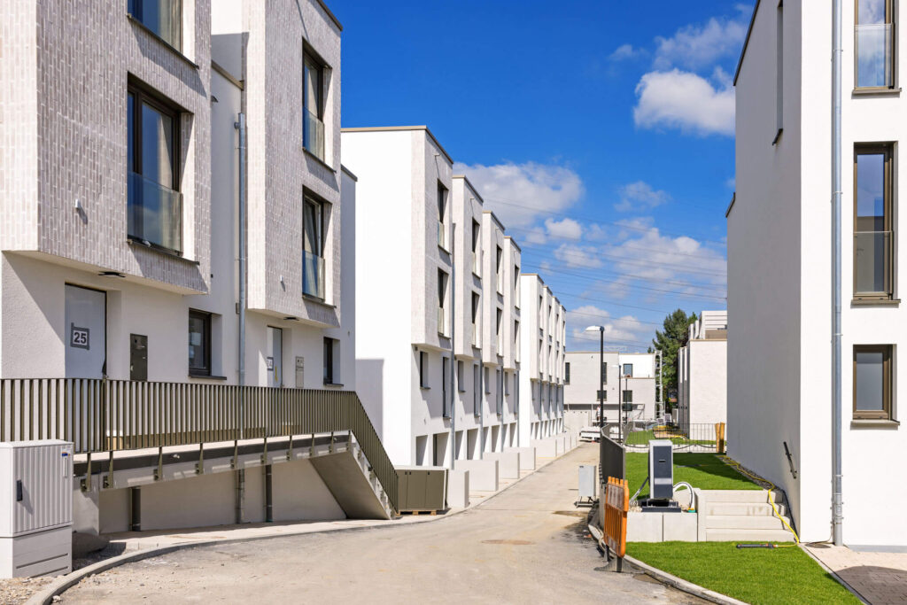 Zehn Einheiten moderner, wunderschöner Reihenhäuser mit gestaffelten Fassaden an einer im Bau befindlichen Straße aufgereiht. An der gegenüberliegenden Straßenseite ein weißes Wohnhaus mit einem gepflegten Grünstreifen davor und einer Baustellenabsperrung davor.