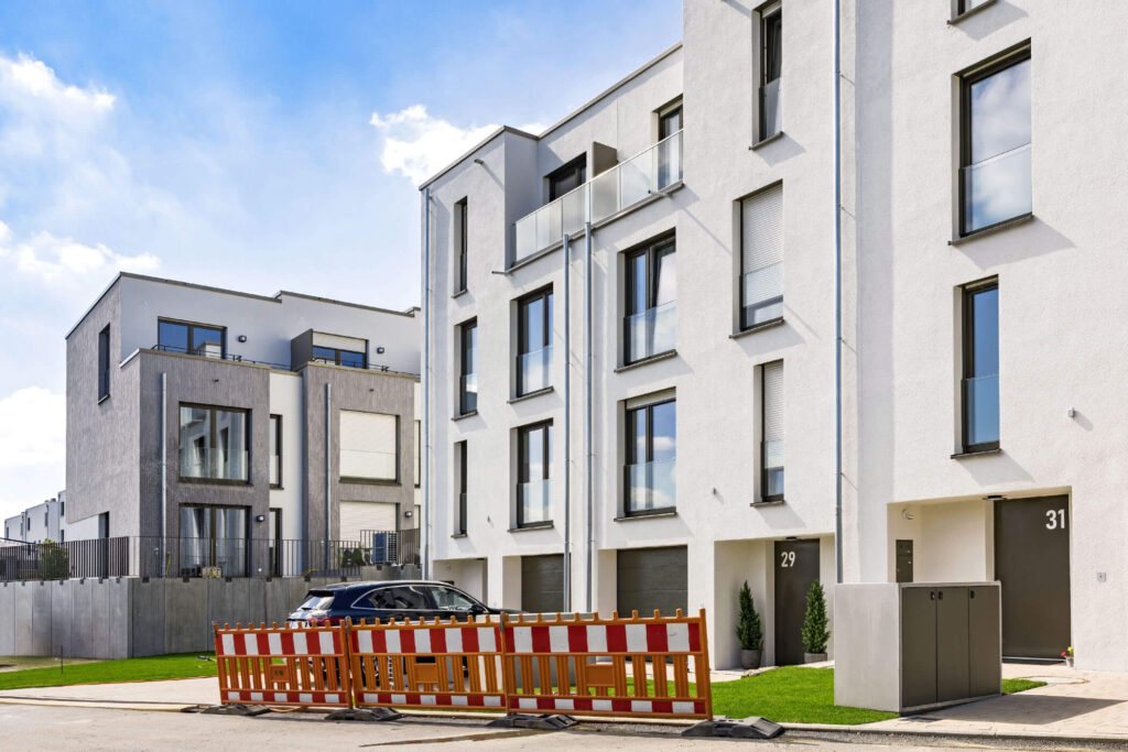 Moderne, wunderschöne Reihenhäuser mit gestaffelten Fassaden, Garageneinfahrten im EG, etwas gepflegtes Grün und Fahrradboxen, an der Einfahrt stehen ein Auto und eine Baustellenabsperrung