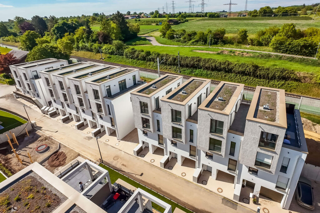 Luftbild eines gehobenen Quartiers aus modernen, wunderschönen Reihenhäusern mit gestaffelten Fassaden und großen Fenstern, Dachterrassen und Dachbegrünung an einer anliegenden Straße. Am Rand der zentrale Quartiersplatz mit Spielplatz im Aufbau, im Hintergrund eine Schallschutzmauer, Schienen und traumhafte Natur mit Feld, Bäumen, Büschen und vereinzelten Strommasten
