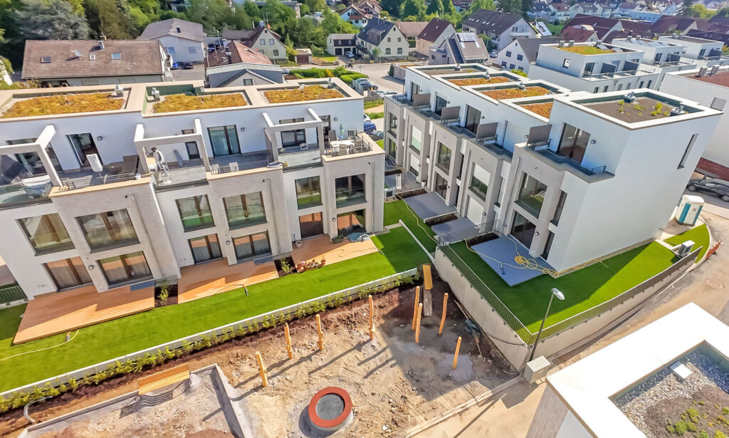 Luftbild eines gehobenen Quartiers aus modernen, wunderschönen Reihenhäusern mit gestaffelten Fassaden und Balkonen, großen Fenstern sowie Terrassen umgeben von großzügigen gepflegten Rasenflächen. Davor ein gemütlicher, zentraler Quartiersplatz im Aufbau mit einem Spielplatz und Bänken.
